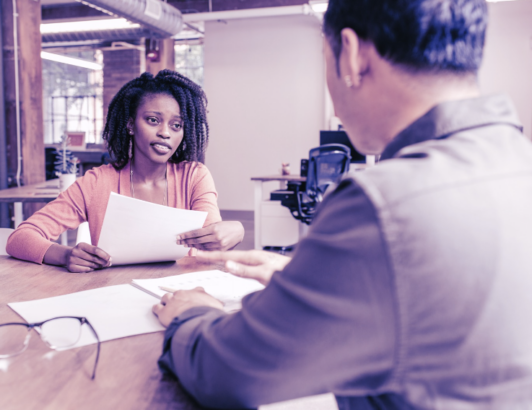 Woman handing document across table during professional interview or review meeting in modern office setting.